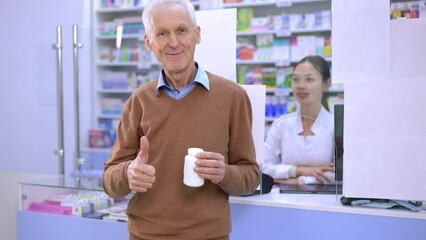 Smiling senior man gesturing thumb up posing with pills in pharmacy with blurred young woman at background. Happy Caucasian client and positive Asian seller in drugstore indoors