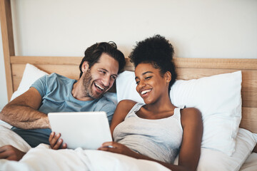 Happy, relaxed and carefree couple reading social media news on digital tablet and laughing in bed. Interracial husband and wife waking up together and browsing internet, sharing a funny online joke - obrazy, fototapety, plakaty