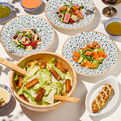 Classic caesar salad with grilled chicken and crouton in wooden bowl on white table. Caesar salad with sauce and wooden spoon. Italian lunch with caesar salad on sunny day. Summer menu.