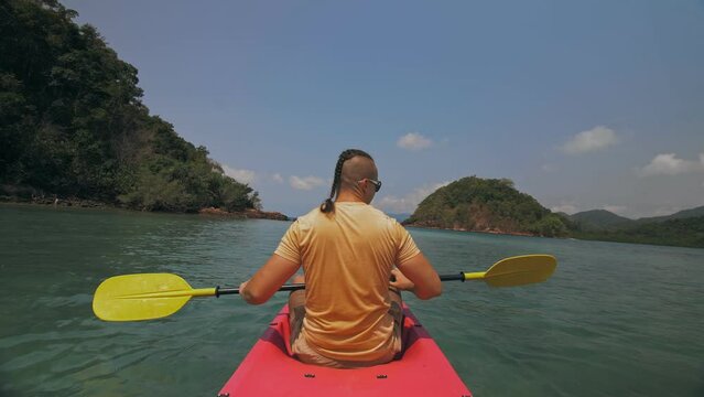 Young man with sunglasses and hat rows pink plastic canoe along sea against green hilly islands with wild jungles. Traveling to tropical countries