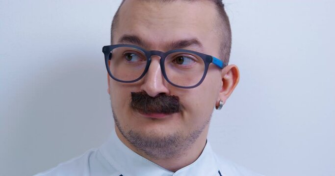 Portrait of young mustachioed man with a fashionable hairstyle, piercing in ear and glasses accusingly raises his eyebrows and purses his lip, looking away at someone, front view