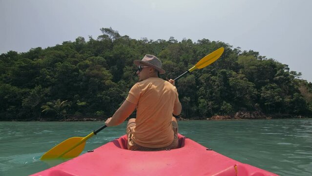 Young man with sunglasses and hat rows pink plastic canoe along sea against green hilly islands with wild jungles. Traveling to tropical countries. Strong guy is sailing on kayak in ocean, back view.