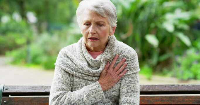 Senior woman suffering from chest pain while sitting outdoors on a park bench. Elderly female having a heart attack outside alone. Mature lady enjoying nature while needing urgent medical attention.