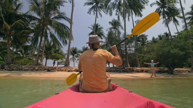 Young man with sunglasses and hat rows pink plastic canoe along sea against green hilly islands with wild jungles. Traveling to tropical countries. Strong guy is sailing on kayak in ocean, back view.