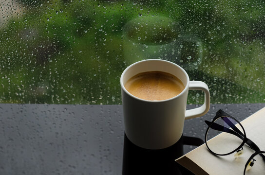 A cup of hot coffee with book and spectacles on table in morning with rain drop on window and green background. Stay home and relaxing concept..