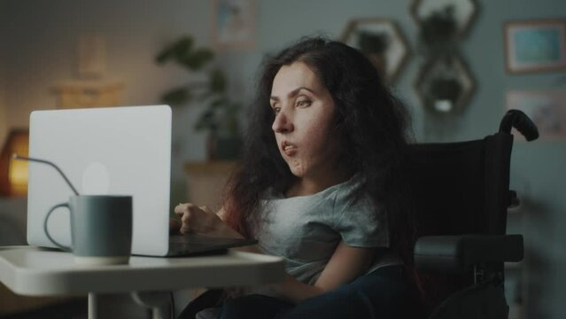 Woman with a disability sitting in a wheelchair at the table, at cozy home and working on a remote job in the internet using laptop
