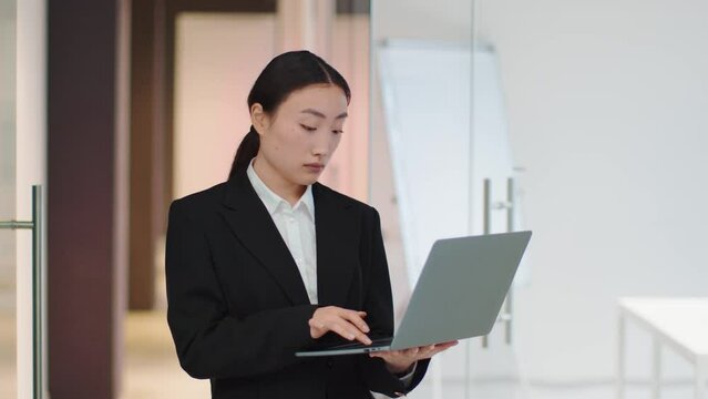 Young asian business woman typing on laptop, answering client, standing at office with computer in hands, free space