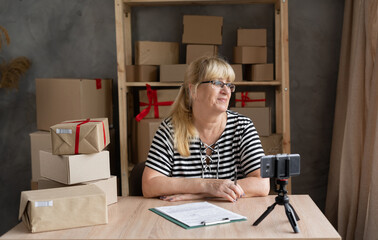 Drop shipping older business owner talking on videocall on mobile phone and taking order. Female entrepreneur working at home office confirming the order on phone. SME telemarketing e-commerce