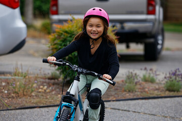 A young girl wearing a pink helmet and black knee pads riding a bicycle with a joyful expression on...