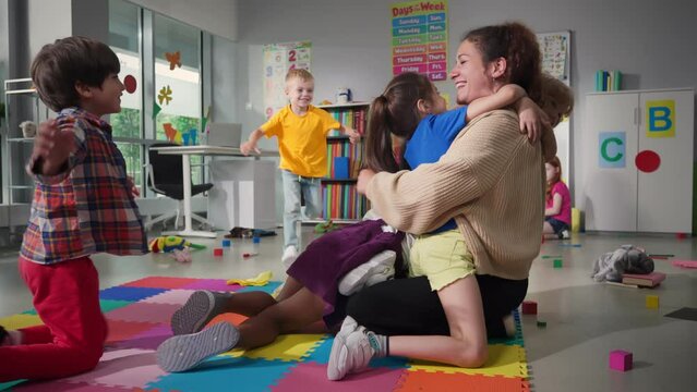 Cheerful smiling teacher and happy diverse schoolchildren hugging in playroom of kindergarten