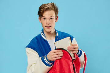 a young, handsome guy student takes out an empty notepad t from his red backpack. Vertical studio photo on a blue background, the concept of preparing for the new study season