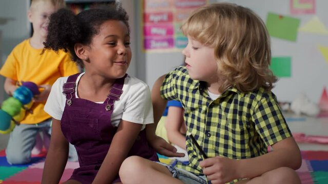 Adorable diverse kids talk and hug sitting on floor in kindergarten playroom