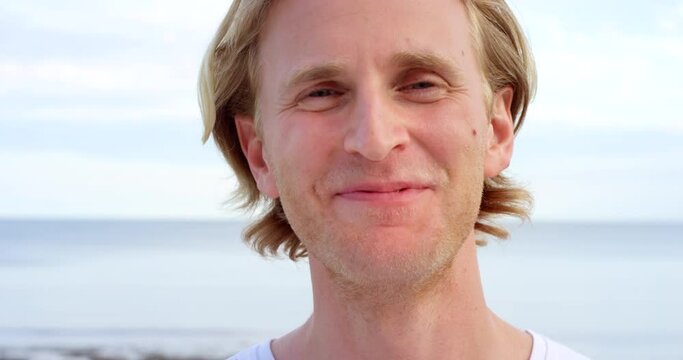 Closeup face portrait of fun and confident young man smiling and laughing with joy at holiday beach getaway. Handsome blonde caucasian male relaxing and standing outside by the sea