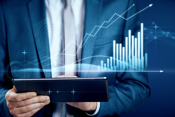 Close up of businessman hand pointing at pad with abstract glowing polygonal business graph hologram with growing arrow on blurry background. Financial growth, market and trade concept.