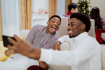 Obraz na płótnie Canvas Elegantly dressed man with afro pulls out phone to take selfie with friend, cousin in shirt, brothers smile, silly funny photos in background family eats Christmas Eve dinner