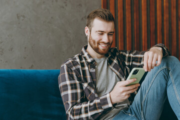 Young man 20s he wear brown shirt hold in hand use mobile cell phone read message chatting sitting on blue sofa in own living room apartment stay home indoor flat on weekends People lifestyle concept