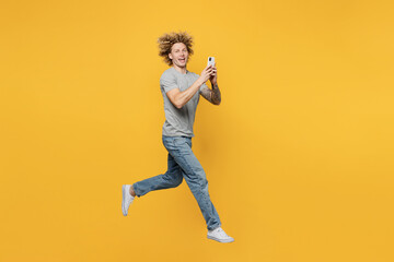 Fototapeta na wymiar Full body side view young caucasian man 20s he wear grey t-shirt look camera jump high hold in hand use mobile cell phone isolated on plain yellow backround studio portrait. People lifestyle concept.
