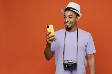 Traveler black man wear purple t-shirt hat hold use mobile cell phone isolated on plain orange color background. Tourist travel abroad on weekends spare time getaway. Air flight trip journey concept.