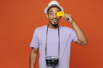 Traveler black man wear purple t-shirt hat hold credit bank card isolated on plain orange color background. Tourist travel abroad on weekends spare time rest getaway. Air flight trip journey concept.