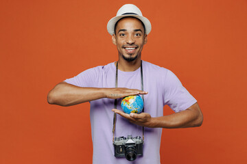 Traveler black man wear purple t-shirt hat hold in palms Earth world globe isolated on plain orange color background. Tourist travel abroad in spare time rest getaway. Air flight trip journey concept.