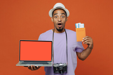 Traveler black man wears purple t-shirt hat hold laptop pc computer passport isolated on plain orange color background Tourist travel abroad in spare time rest getaway Air flight trip journey concept.
