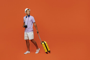 Full size traveler black man wear purple t-shirt hat headphones go with suitcase isolated on plain orange background. Tourist travel abroad in spare time rest getaway. Air flight trip journey concept.