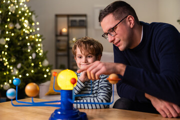 Fototapeta na wymiar Ambitious man spends time with school-age son during Christmas holidays, dad teaches child about planetary system, they assembled toy model together, guy points to sun
