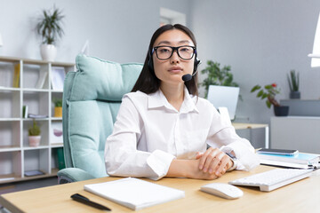 Portrait of a young beautiful Asian woman in glasses and headphones with a microphone, sitting in the office at the desk conducting an online webinar. He looks at the camera, smiles.