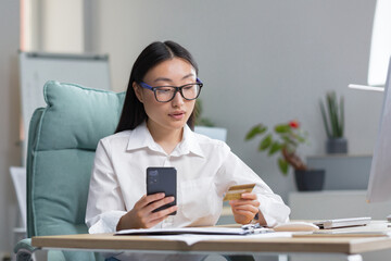 Online shopping. A young beautiful Asian woman holds a mobile phone and a credit card in her hand, makes an order on the phone, pays by card.. Sitting at a table in a modern office.