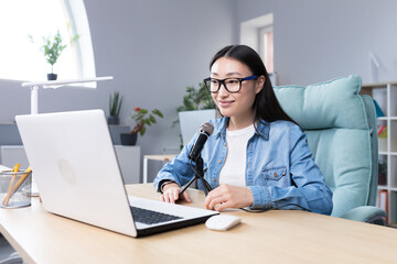 Podcast recording, interview. Young beautiful Asian woman sitting at a table with a microphone and a laptop. Talks, tells. Makes an audio recording of a podcast, gives an interview via video call.