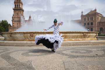Young teenage woman in white dance suit with black polka dots and green carnations in her hair, dancing flamenco in front of a water fountain. Flamenco concept, dance, art, typical Spanish dance. - obrazy, fototapety, plakaty