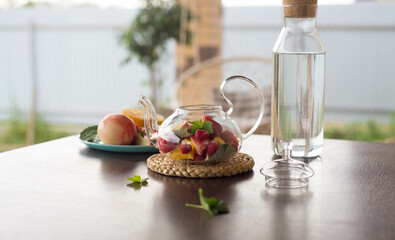 A glass teapot with fruits and berries, mint leaves, a bottle of water on the table outside. Summer fruit tea.