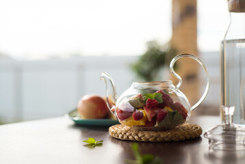 A glass teapot with fruits and berries, mint leaves, a bottle of water on the table outside. Summer fruit tea.