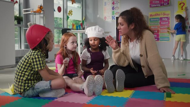 Kids dressed as firefighter, doctor and cook sit on carpet and listen to teacher in kindergarten
