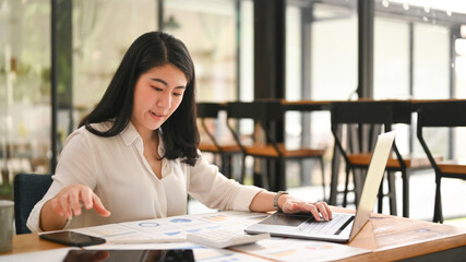 Professional business woman examining the numerical data on financial document and using laptop computer at office desk