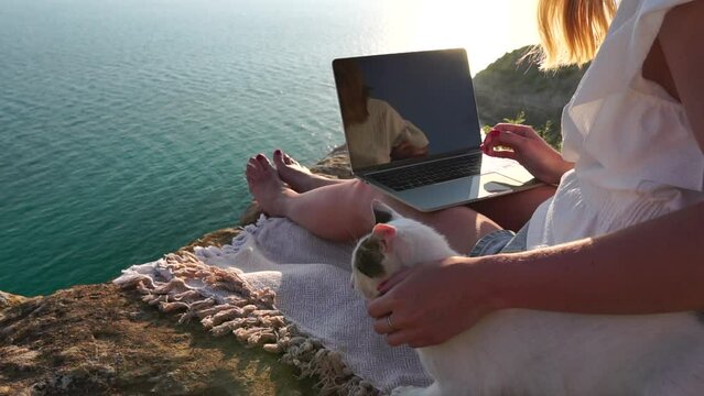 Successful business woman in yellow hat working on laptop by the sea at sunset time. Pretty lady typing on computer at summer day outdoors. Freelance, digital nomad, travel and holidays concept.