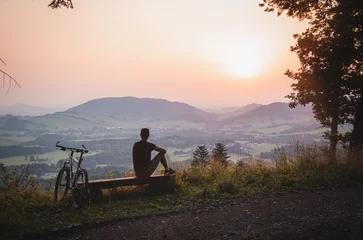 Tuinposter Active athlete sitting on a bench after going to the top of a hill on his bike and watching the sunset. Beskydy mountains, Czech republic, heart of Europe © Fauren