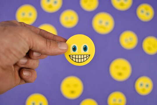 Closeup shot of hand holding happy smiling emoji face between sad emoj faces on purple background 