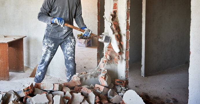 Close up of sledgehammer blow on brick, plaster. Workman striking devastating blow at remnants of interoom wall against backdrop of table and other plastered walls.