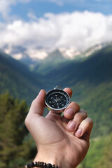 A hand with a compass against the backdrop of epic snow-capped mountains with clouds and a forest at the foot, close-up