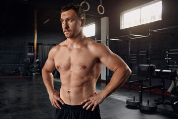 Fototapeta na wymiar Portrait Of Handsome Muscular Man Posing In Gym. Confident Fitness Trainer Standing In Modern Sport Club Interior And Looking Away. Advertising Healthy Lifestyle 