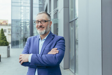 Portrait of successful happy and satisfied investor, businessman in glasses and business suit outside office building, banker looking at camera and smiling, financier with crossed arms