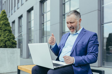 Successful gray haired businessman working on laptop celebrating victory triumph overweight man sitting on bench outside office building