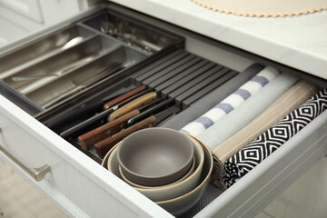 Open drawer of kitchen cabinet with different utensils, dishware and towels, closeup