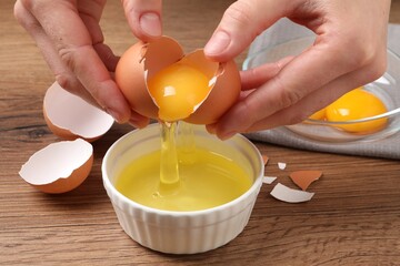 Woman separating egg yolk from white over bowl at wooden table, closeup