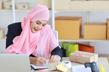 Active asian woman in muslim suit sitting and working with computer, mobile smartphone and online package box delivery. Startup small business freelance working girl checking customer order from book