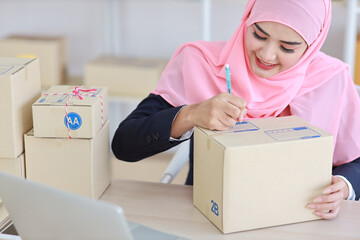 Fototapeta Active smiling asian woman in blue muslim suit sitting and working with online package box delivery. Startup small business SME freelance girl working on computer and mobile phone with happy face. obraz