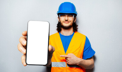 Studio portrait of smiling man, construction worker, holding big smartphone with blank on screen in...