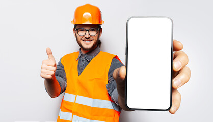 Studio portrait of young smiling construction worker, holding big smartphone with blank on screen...