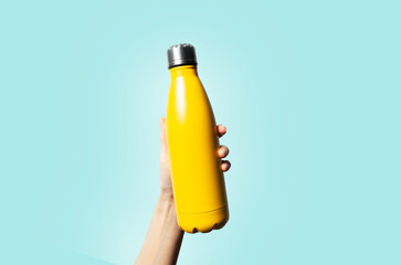 Close-up of female hand holding reusable water bottle of yellow color on cyan background.